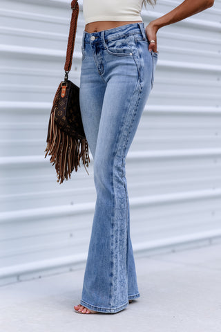 A person wearing light-wash blue flare jeans, standing barefoot against a white corrugated metal wall and holding a brown fringe handbag.