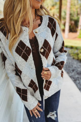 A woman wearing a light gray and dark brown argyle knit cardigan over a dark brown top, paired with blue jeans, standing outdoors.