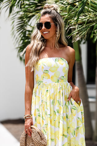 Woman wearing a yellow strapless dress with a lemon print, standing outdoors with greenery in the background.