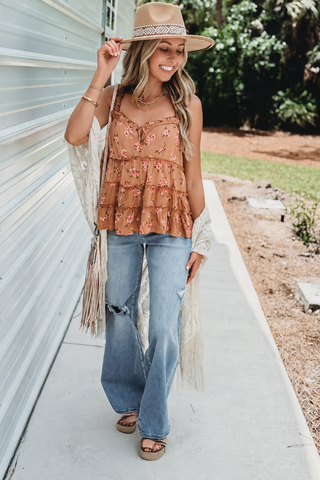 A woman stands on a concrete walkway wearing a bohemian-style outfit featuring a rust-colored floral peplum top, distressed light-wash jeans, a cream fringe cardigan, a tan hat, and brown sandals.