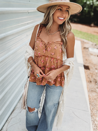 A woman wearing a bohemian-style outfit featuring a rust-brown floral tiered tank top, a white fringed cardigan, distressed blue jeans, a wide-brim straw hat, and gold jewelry, standing outdoors next to a white corrugated metal wall.
