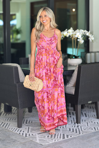 A woman stands in a modern patio setting wearing a bright pink floral maxi dress with orange and peach tones, holding a woven straw clutch bag.