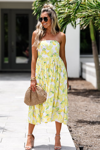 Woman in a yellow strapless dress with lemon pattern holding a straw bag outdoors.