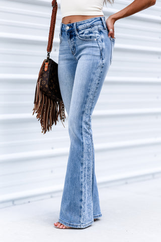 A person wearing light-wash blue flare jeans, shown from the waist down, holding a brown fringe shoulder bag against a white corrugated metal wall.