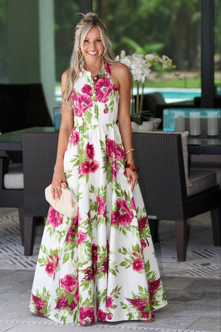 A woman stands in a patio setting wearing a sleeveless white halter maxi dress with a bold pink and green floral print. She is holding a small beige clutch.