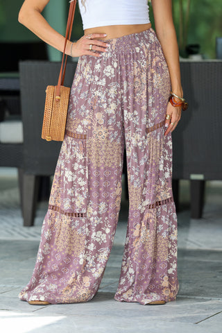A person wearing loose, wide-leg boho pants in a dusty mauve-purple color with a beige and white patchwork floral print. The pants feature a high waist, elastic waistband, and tiered ruffle details.