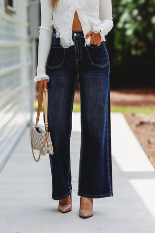 A person wearing dark blue wide-leg denim jeans, a white lace-trim blouse, and beige heels, holding a small patterned handbag on a sidewalk.