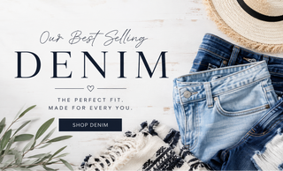 A styled flat-lay featuring folded pairs of blue denim jeans on a white wood background, with a straw hat, cotton stems, and a black-and-white fringed throw. Large text reads 'Our Best Selling DENIM' with a 'SHOP DENIM' button.