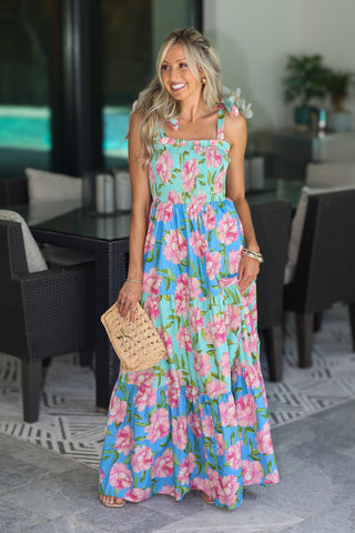 A woman wearing a vibrant, multicolor floral maxi dress with pink flowers and green leaves on a light blue background, holding a woven straw clutch in an outdoor patio setting.