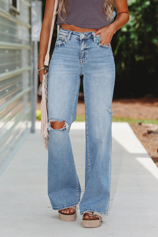 A person wearing light-wash blue wide-leg distressed jeans with ripped knees, paired with a dark gray crop top and platform sandals, standing outdoors on a concrete walkway.