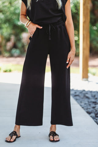 A person wearing black wide-leg cropped pants with a drawstring waist, paired with a black sleeveless top and black sandals, standing outdoors on a concrete path with blurred greenery in the background.