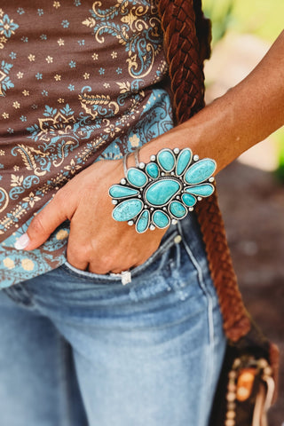 A close-up lifestyle shot of a person wearing a large, statement-style cuff bracelet with multiple turquoise-blue stones set in silver. The person is also wearing a brown paisley top, blue jeans, and a brown braided shoulder bag.