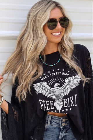 A woman wearing a black sheer lace open-front cardigan over a black graphic T-shirt with an eagle design and the words 'FREE BIRD', paired with denim shorts and sunglasses, posing against a white paneled wall.