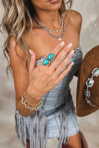 A woman wearing a western-style denim outfit and red cowboy boots, styled with layered silver chain necklaces and bracelets, a turquoise ring, and a brown western hat. She is posing against a neutral textured gray background.