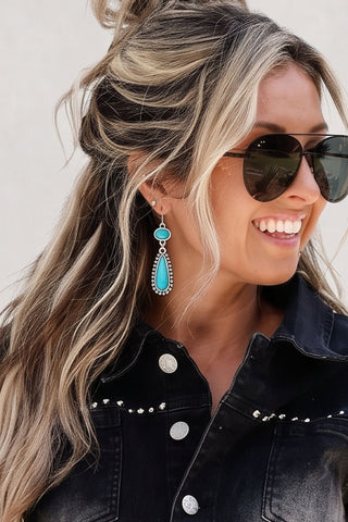 A woman modeling a black denim short-sleeve button-up shirt and matching denim shorts with western-style silver buckles and studs, styled with sunglasses, turquoise earrings, a small handbag, and black boots against a light gray wall.