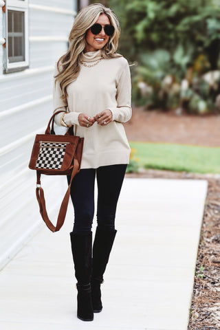 Woman wearing a cream turtleneck sweater, black leggings, and knee-high black boots, holding a checkered handbag.